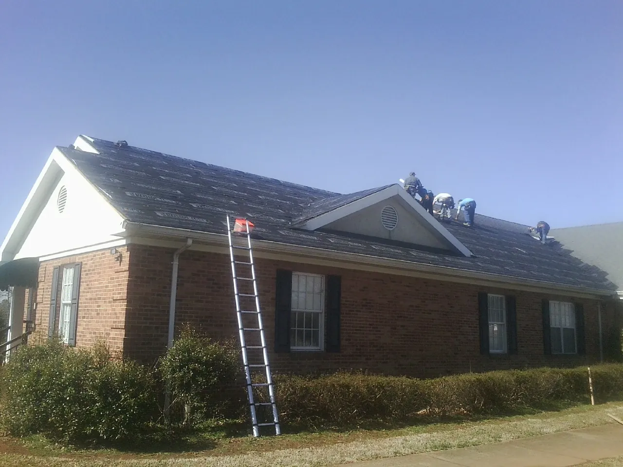 Crew tearing shingles off a brick ranch roof with a ladder set — Emergency Roof Repair work by our crew in Hays