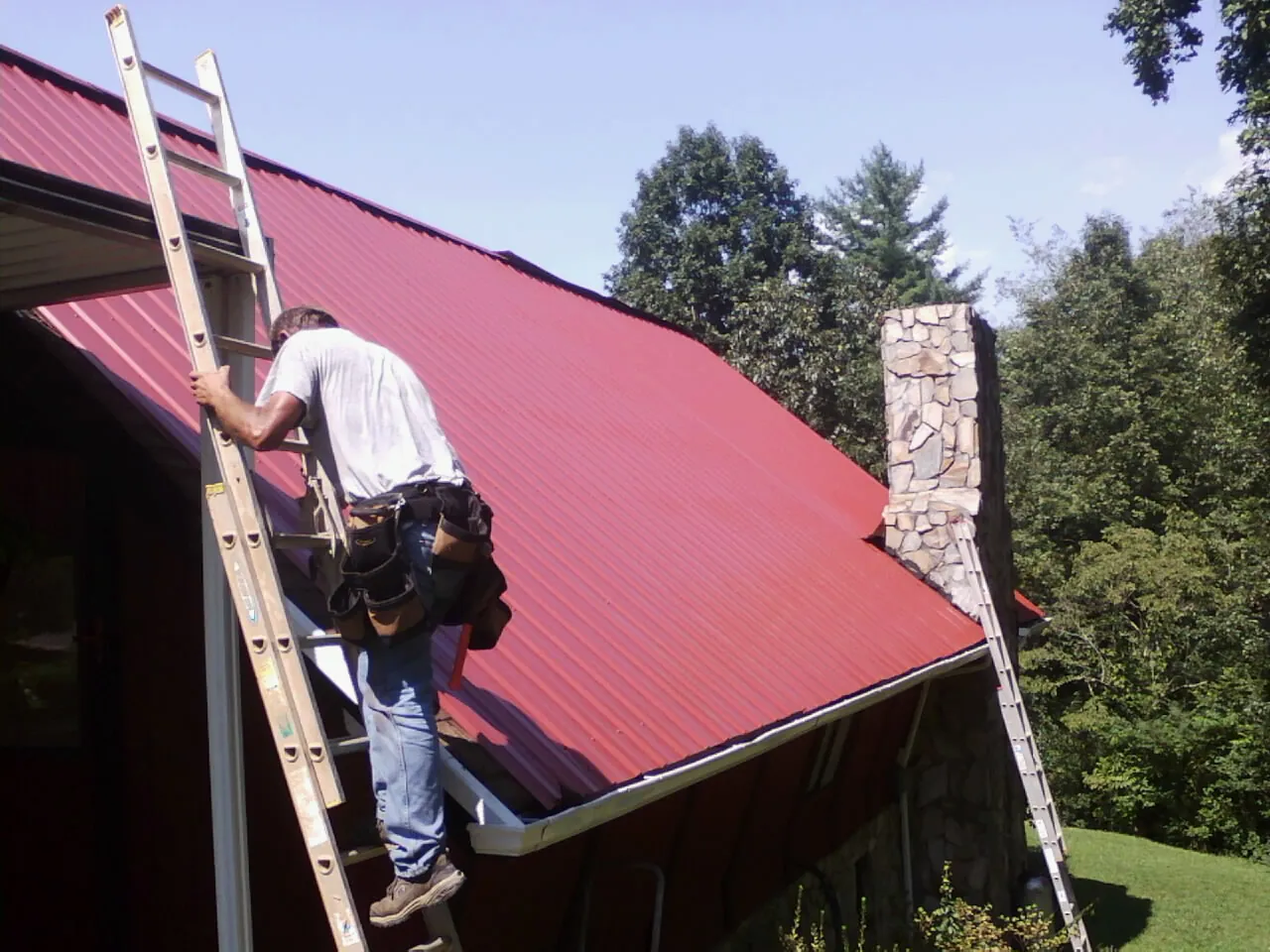 Roofer on a ladder beside a red metal roof and stone chimney — Soffit & Fascia Repair by Roofing Brothers in Hays